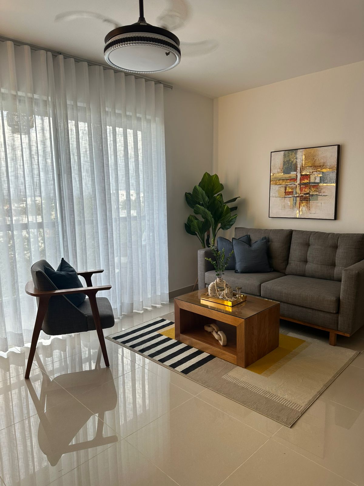 A stylish living area is presented, featuring a cozy gray sofa complemented by two decorative pillows. A wooden coffee table sits atop a patterned rug, enhancing the space. A large window, dressed with sheer curtains, allows natural light to fill the room, while a potted plant adds a touch of greenery.