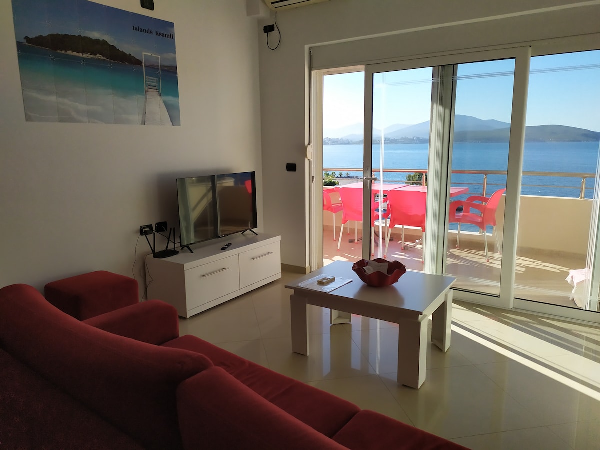 A bright living area showcases a flat-screen TV and a white coffee table, complemented by a red sofa. Large glass doors provide a view of the sea and mountains, leading to a balcony furnished with red chairs and a table.