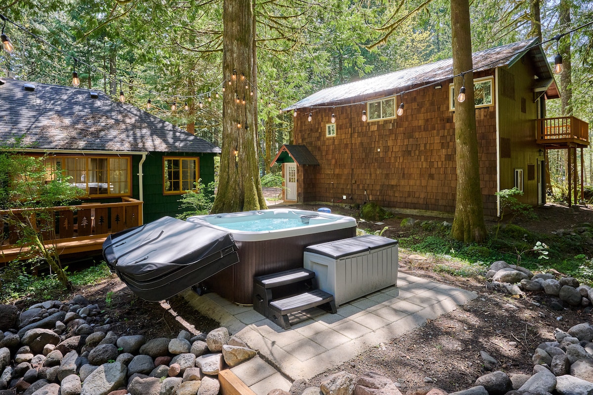 An inviting outdoor area features a 7-person hot tub surrounded by a stone patio, nestled among tall trees. Two cabins are visible in the background, showcasing natural wood siding and warm tones, with string lights elegantly draping overhead, enhancing the serene setting.
