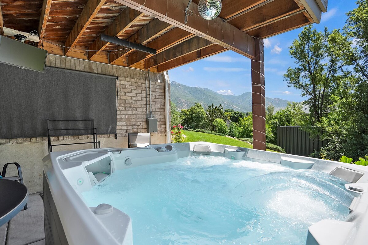 A hot tub is situated on a covered patio, surrounded by natural greenery and mountain views. Sunlight filters through, creating a serene atmosphere. The space includes a few outdoor chairs, providing a relaxing setting to enjoy the surroundings.
