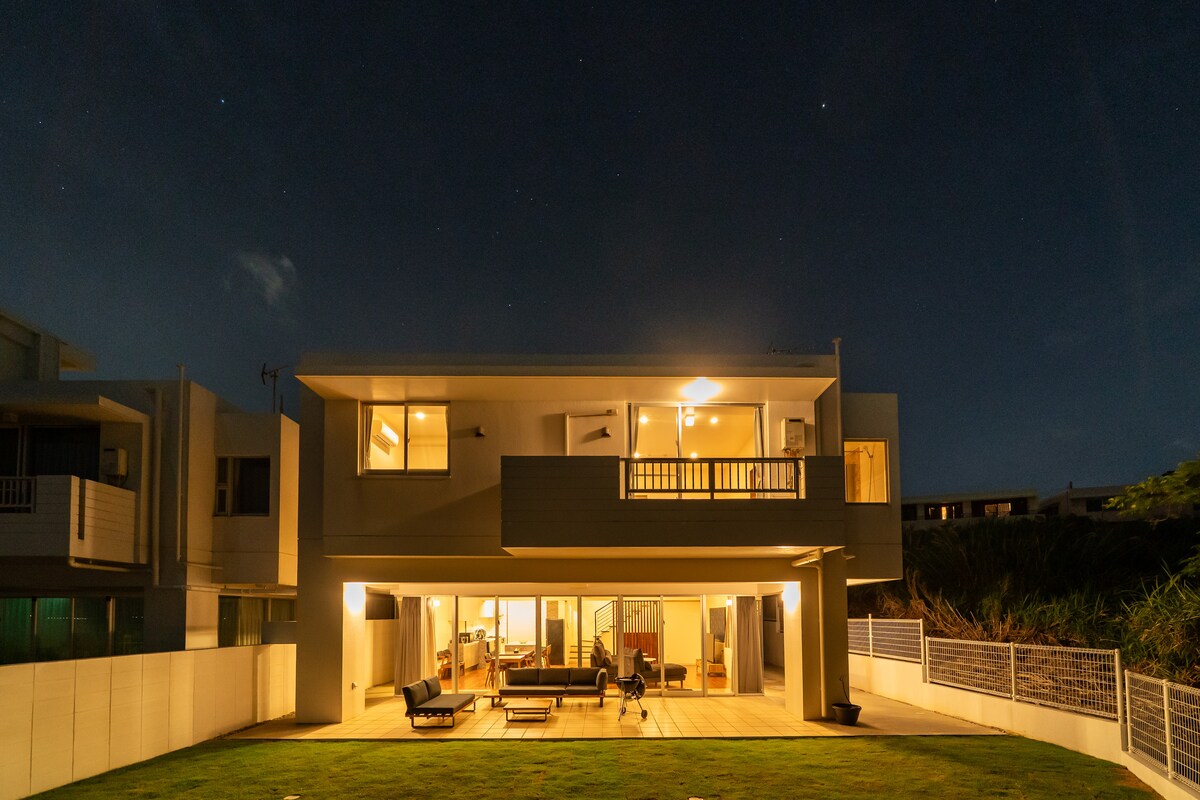 The exterior of a two-story house is illuminated against the night sky, showcasing large glass doors that provide a view of the interior living area. Outdoor furniture is arranged on a spacious deck, surrounded by a well-maintained lawn.