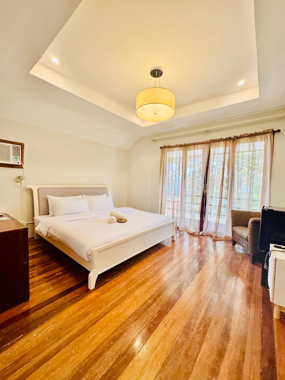 A bright bedroom features a spacious bed with crisp white linens, complemented by a cozy armchair and a wooden bedside table. Large windows allow natural light to fill the room, while warm wooden floors enhance the inviting atmosphere.