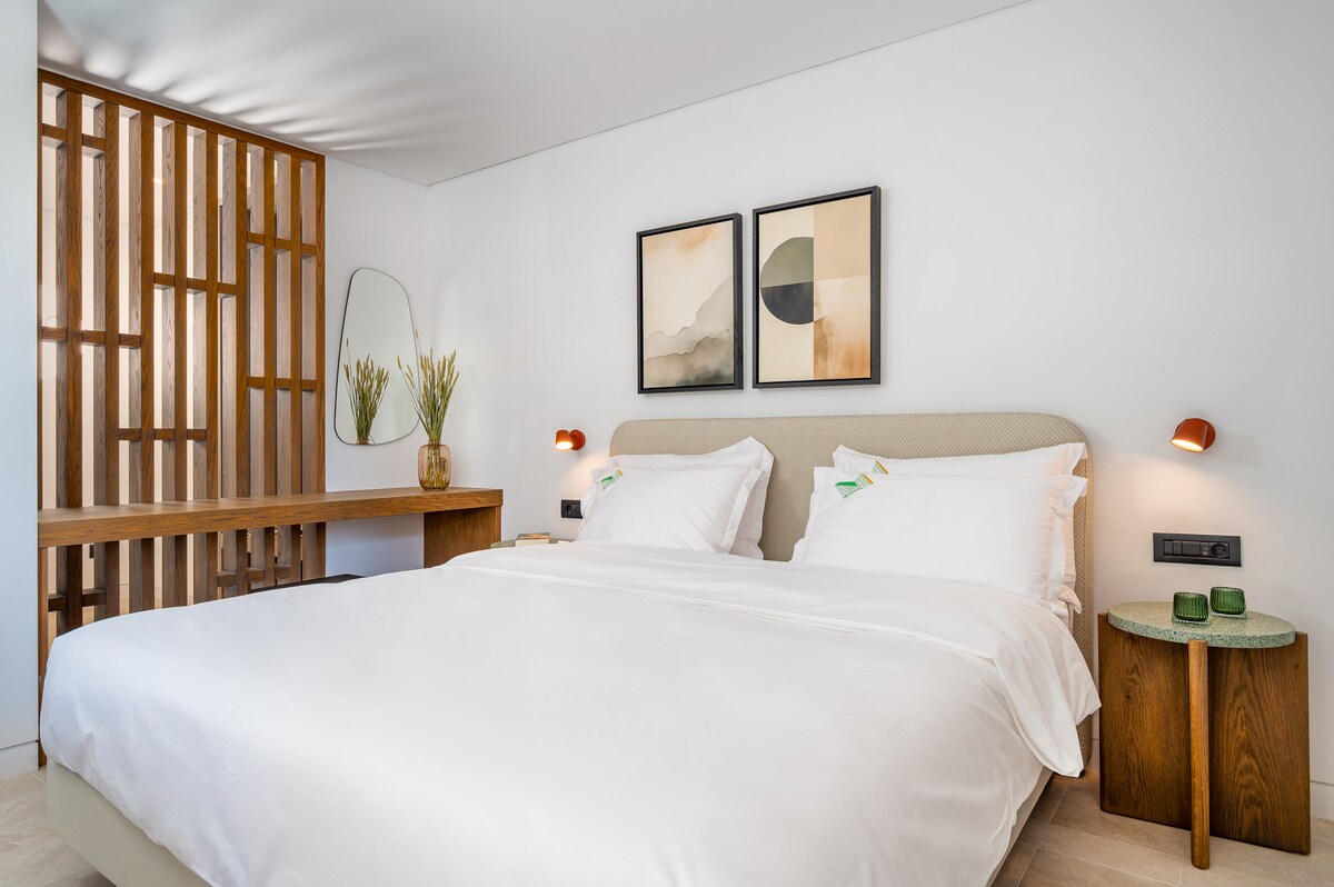 A well-appointed sleeping area features a spacious bed with crisp white linens. Two framed artworks decorate the wall above the headboard. A wooden side table and a mirror reflect natural light, while a green accent table adds a pop of color near a minimalist workspace.