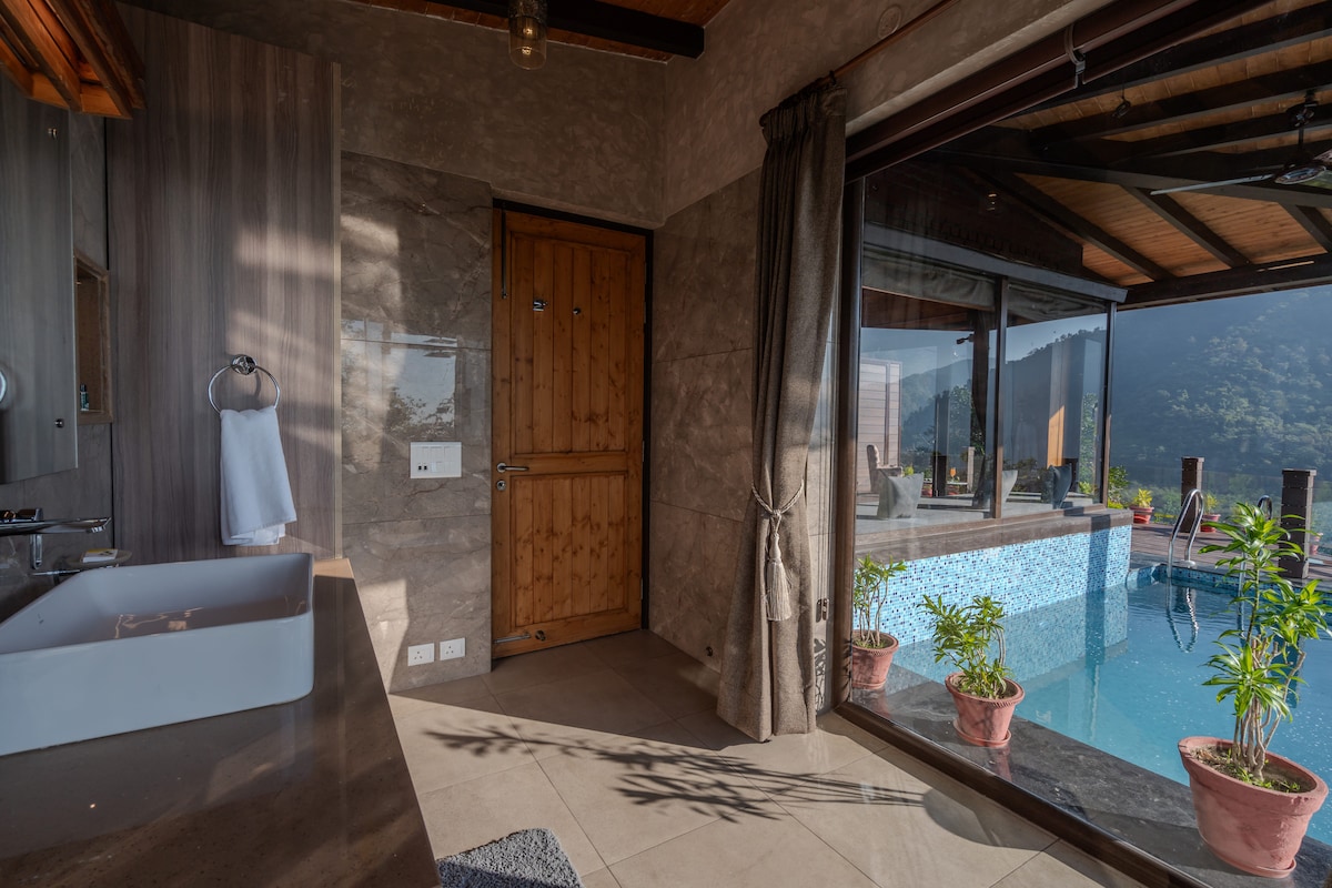 An elegantly designed bathroom features natural stone walls and a large soaking tub. A door opens to a private area with a shimmering pool and lush plants, framed by expansive glass panels that invite natural light and showcase scenic mountain views.