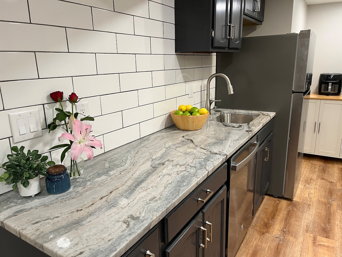 A modern kitchenette features polished granite countertops with a bowl of fresh citrus fruits displayed. A stainless steel sink and dishwasher are situated beside a full-size refrigerator. A bouquet of fresh flowers adds a touch of color to the space, accompanied by small potted plants.