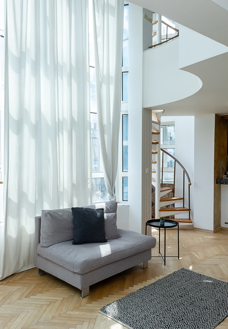 A bright living area is featured, showcasing a comfortable gray couch complemented by a dark cushion. Natural light floods the space through tall windows, with sheer white curtains gently flowing. A small black side table is positioned next to the couch on a patterned rug.