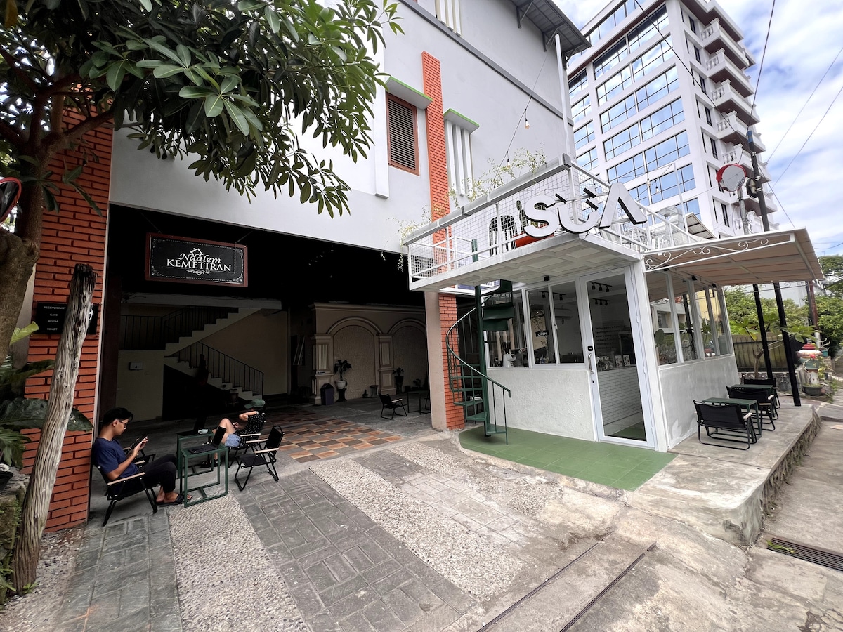 The exterior of the property is displayed, featuring a modern building with a welcoming entrance. Outdoor seating areas with black chairs are positioned along the pathway, surrounded by greenery. A spiral staircase is visible, leading to an upper level, while a sign for Ndalem Kemetiran is prominently featured.