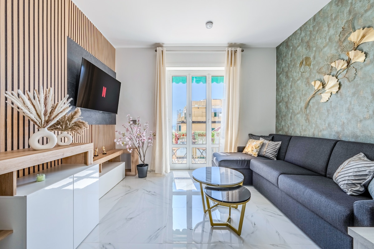 A modern living room features a comfortable gray sofa arranged opposite a sleek entertainment unit with a mounted TV. Natural light enters through large, open doors leading to a balcony. Decorative elements include wall art and a small indoor plant.