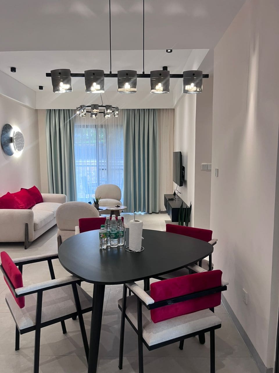 A dining area features a black circular table surrounded by four chairs with a contemporary design. Above, overhead lighting provides illumination. A cozy living area is visible in the background, highlighted by a plush sofa adorned with bright cushions and sheer curtains that frame the window.