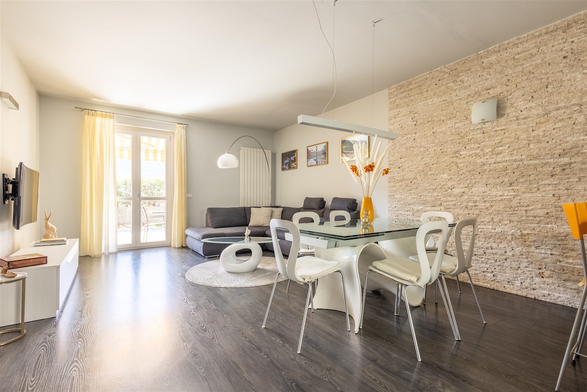 A spacious living area features a modern glass dining table surrounded by white chairs, complemented by a cozy sofa in the background. Natural light enters through large windows with light curtains, illuminating the tasteful decor of the room, including a stone accent wall.