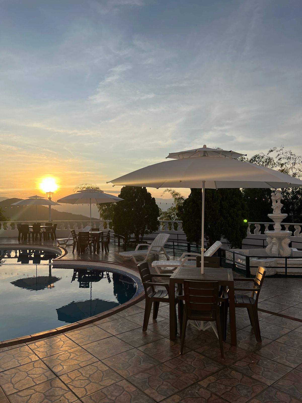 A tranquil outdoor space is depicted, featuring a swimming pool surrounded by tables and chairs. Two umbrellas provide shade as the sun sets on the horizon, casting a warm glow over the area. Lush greenery is positioned around the pool, enhancing the serene environment.