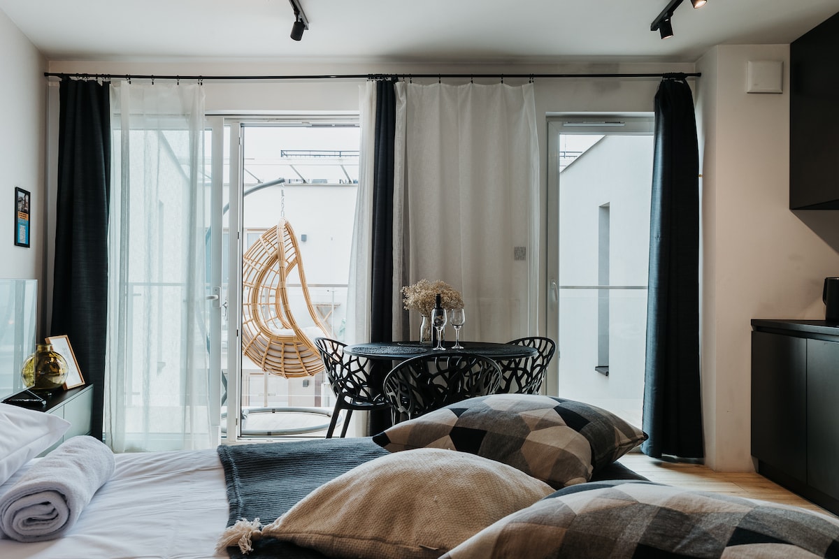 A cozy room showcases a spacious bed adorned with plush pillows and a soft blanket. Large glass doors open to a private balcony featuring a hanging chair and a small dining table. Sheer curtains allow natural light to brighten the interior.