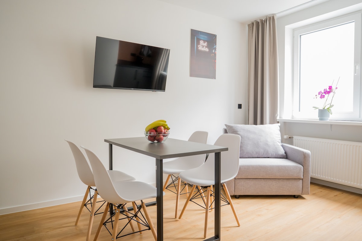 A dining area features a square table surrounded by four white chairs, with a bowl of fresh fruits placed on the table. A compact gray sofa complements the space. A wall-mounted television and a window with light curtains enhance the bright atmosphere.