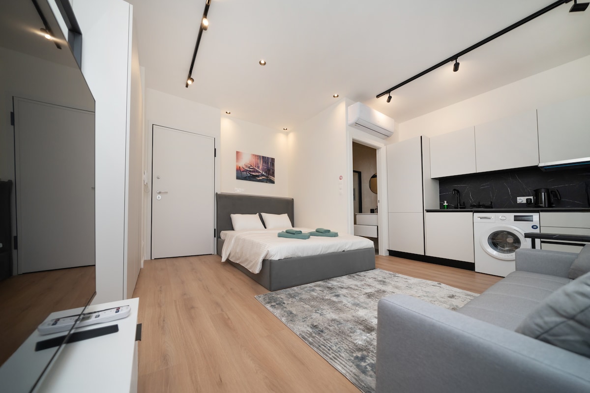 A modern studio features a spacious bed adorned with soft linens and towels. A sleek kitchenette is visible, along with a washing machine. Natural light filters through the room, highlighting the light wood flooring and a comfortable seating area with a neutral-colored sofa.