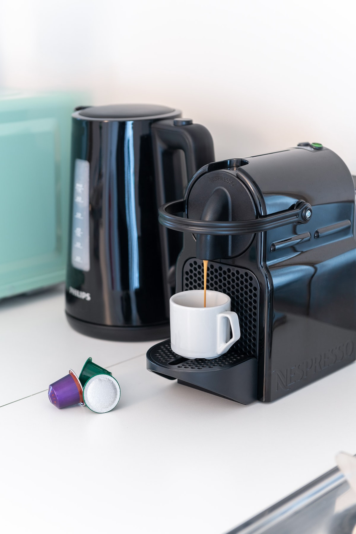 A Nespresso coffee machine is positioned on a countertop, dispensing coffee into a white cup. A kettle is nearby, and colorful coffee capsules can be seen resting on the surface.