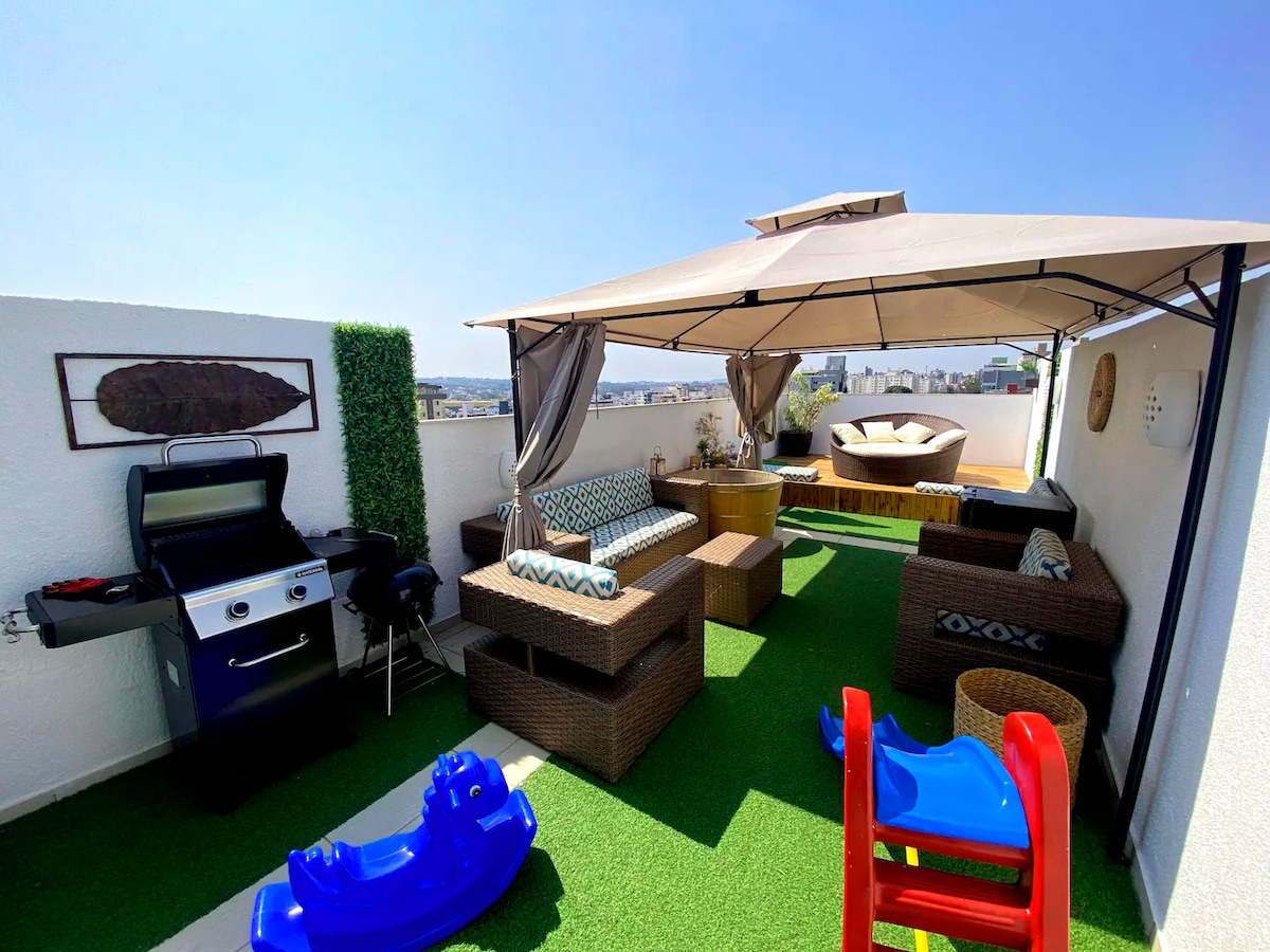 An outdoor terrace is covered by a large beige canopy, providing shade over various seating areas. Comfortable wicker furniture is arranged around a central coffee table. A grill is positioned nearby, alongside a playful red and blue slide, with artificial grass creating a vibrant atmosphere.