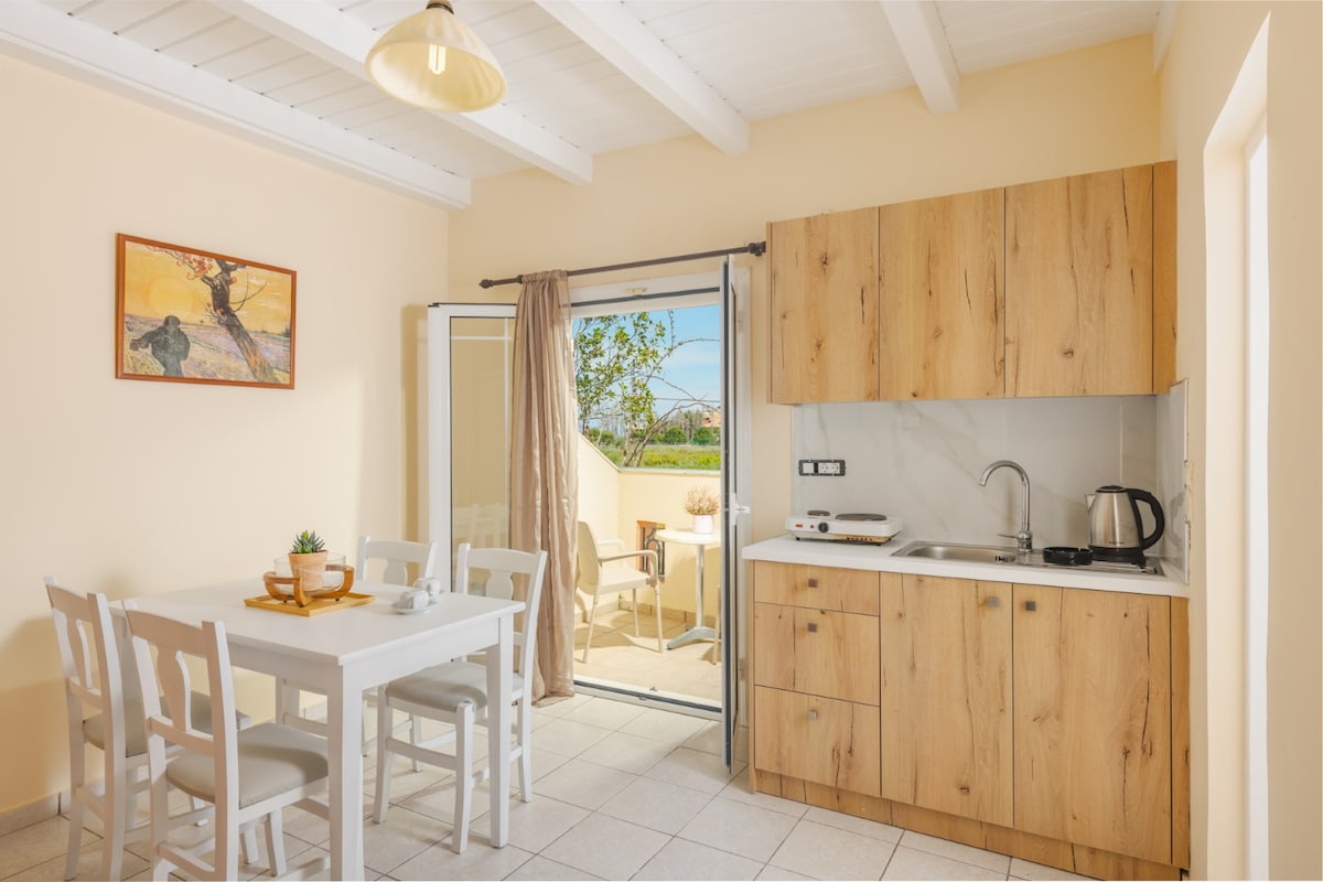 A bright dining area features a white table surrounded by four chairs, complemented by natural wood cabinetry in the kitchenette. Glass doors open to a small balcony, allowing for garden views and sunlight to fill the space. A piece of artwork adorns the wall.