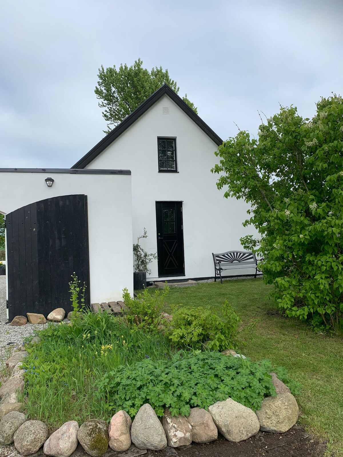 The exterior of the annex is presented, featuring a white façade with a black door and window accents. A bench is positioned on a stone path, surrounded by greenery and landscaped rocks. The entrance is framed by a decorative gate and low shrubs.