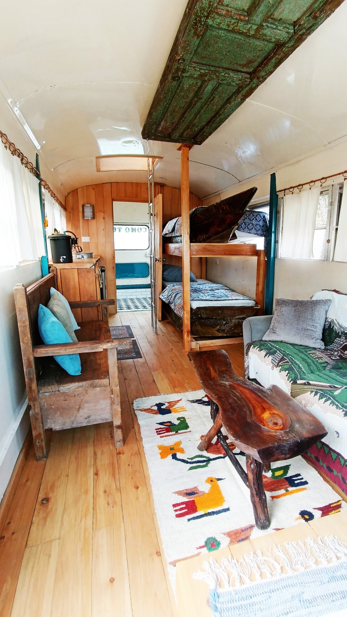 The interior of the chicken bus features wooden flooring and unique bunk beds crafted from recycled canoes. A cozy living area includes a bench with cushions, while a decorative rug adds color to the space. Natural light brightens the room through multiple windows.
