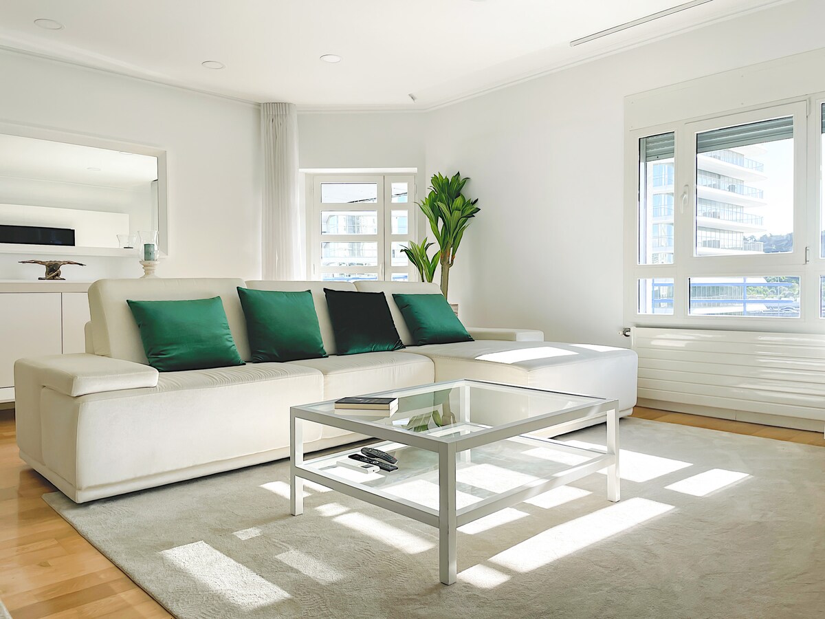 A spacious living area is filled with natural light from large windows. A plush white sofa is adorned with green cushions, accompanied by a glass coffee table. A plant adds a touch of greenery, enhancing the calm and stylish vibe of the room.