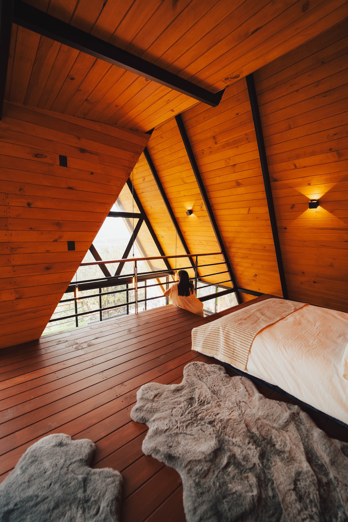 A spacious loft area features warm wooden walls and ceiling, with large triangular windows allowing natural light. A cozy bed is positioned on polished wooden flooring, accompanied by soft, textured rugs. Wall sconces provide gentle illumination, enhancing the serene ambiance of the space.