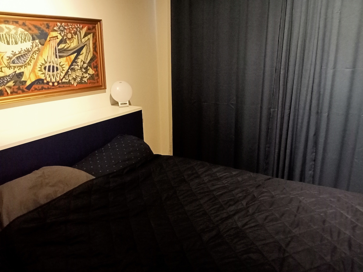 A cozy sleeping area features a spacious bed with a dark comforter and pillows. A nightstand holding a small lamp is positioned beside the bed. Dark curtains cover the window, and a piece of wall art adds a touch of color to the warm atmosphere.