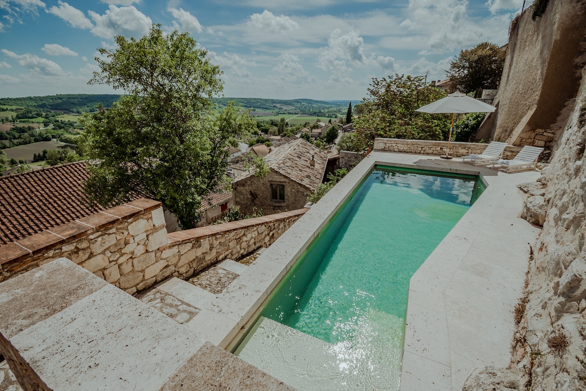 A serene stone pool is positioned against a scenic backdrop of rolling hills and a clear sky. Surrounding the pool, natural stone walls provide an elegant structure, while a shaded area is created by a large umbrella. Green trees and terracotta roofs can be seen in the distance.