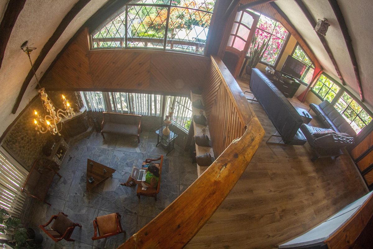 The open living area features vaulted ceilings with wooden beams. Natural light flows through large windows, illuminating the comfortable seating arrangements and decorative plants. A polished stone floor and wooden accents enhance the inviting atmosphere for relaxation and connection.