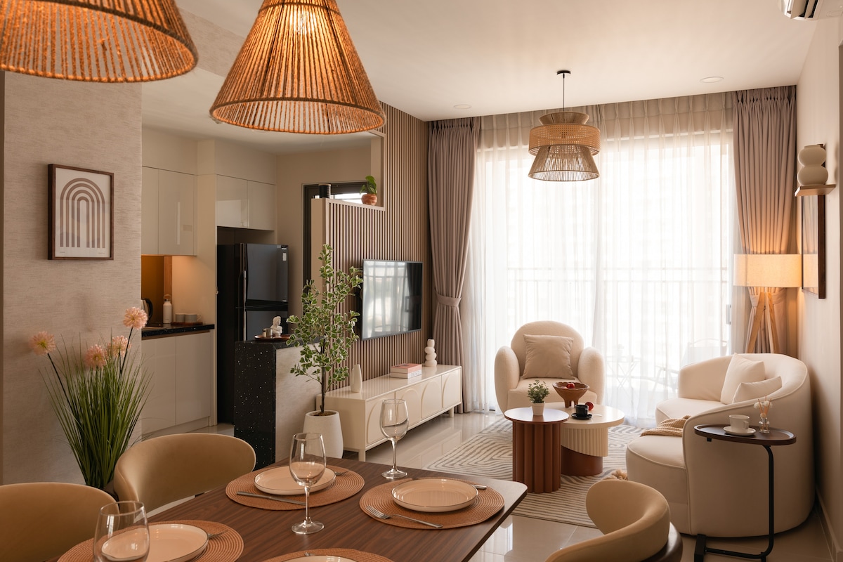 An open-plan living area features a cozy seating arrangement with light-colored furniture, complemented by natural light streaming through large, sheer windows. A dining table is set for six, with elegant place settings. Modern lighting fixtures hang above, adding to the stylish ambiance.