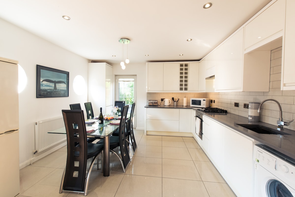 A modern kitchen is equipped with sleek white cabinetry and stainless steel appliances. A rectangular dining table is surrounded by six black chairs. Natural light pours in through large windows, illuminating the polished tile flooring.