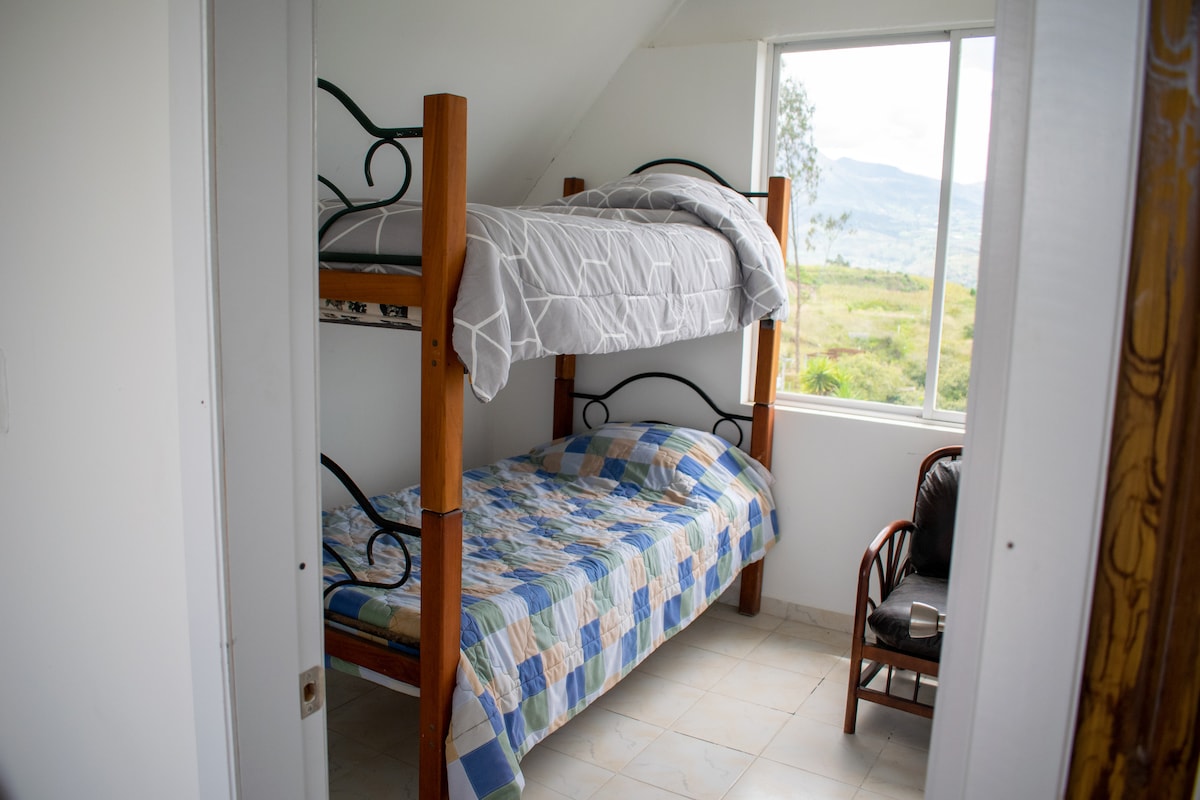 A well-lit room features a wooden bunk bed, with a twin-sized mattress on the top and a larger mattress below. A patterned quilt covers the beds. A chair and a window providing natural light overlook a scenic landscape.