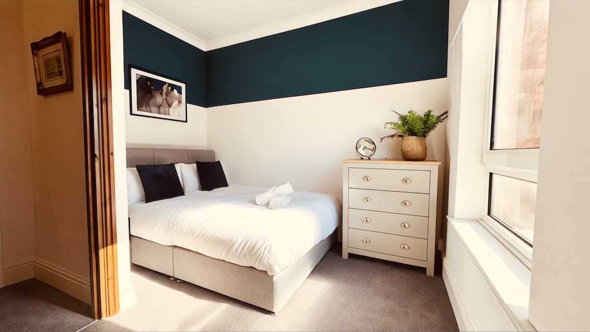 A calming double bedroom is featured with a spacious bed dressed in crisp white linens. A light-colored dresser stands against the wall, complemented by a decorative plant and a clock on its surface, while natural light streams through a large window.