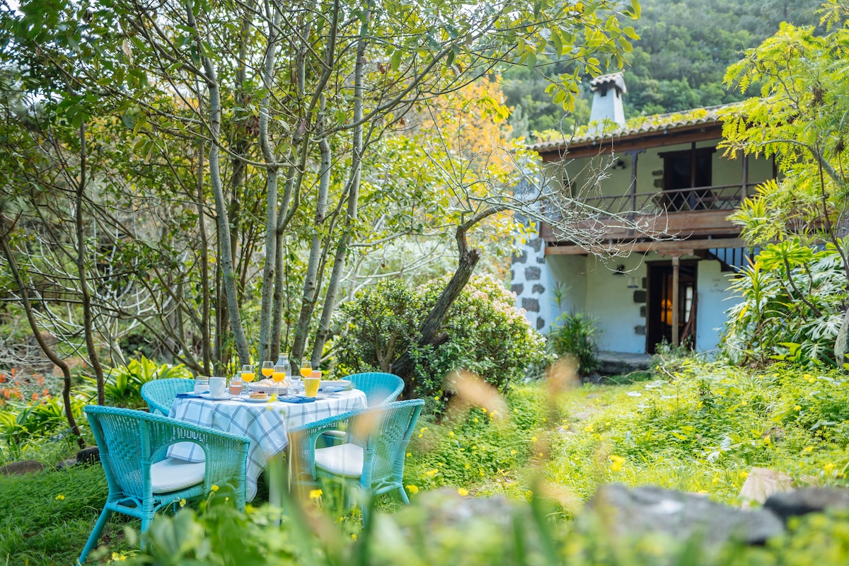 A charming outdoor dining area is surrounded by lush greenery and vibrant flowers. A table set with drinks is flanked by four woven chairs. The rustic house with a balcony and a stone exterior is visible behind the trees, creating a serene and inviting setting.