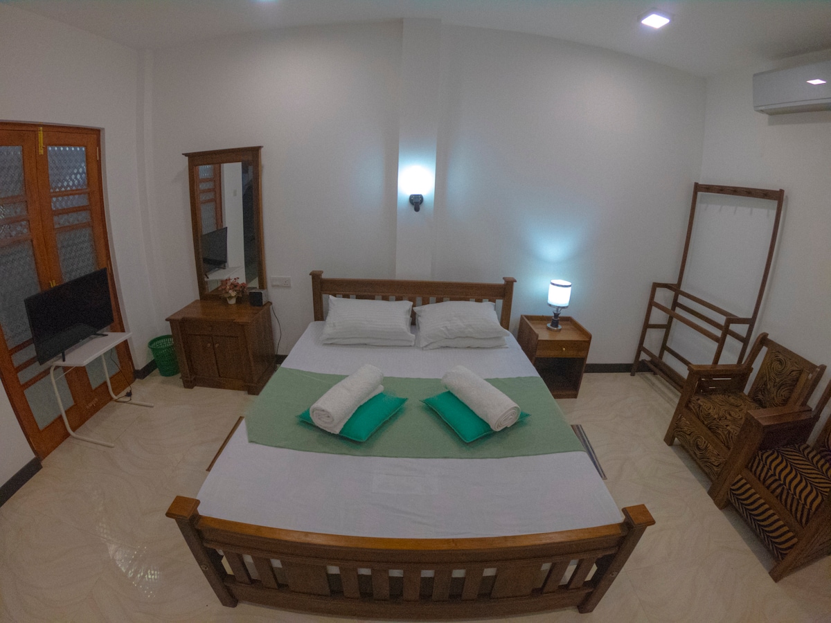 A spacious guest room is presented with a comfortable bed, topped with fresh towels. A wooden dresser and a chair are visible, alongside a flat-screen television. Soft lighting from a bedside lamp creates a calming atmosphere, complemented by a neatly arranged area for relaxation.