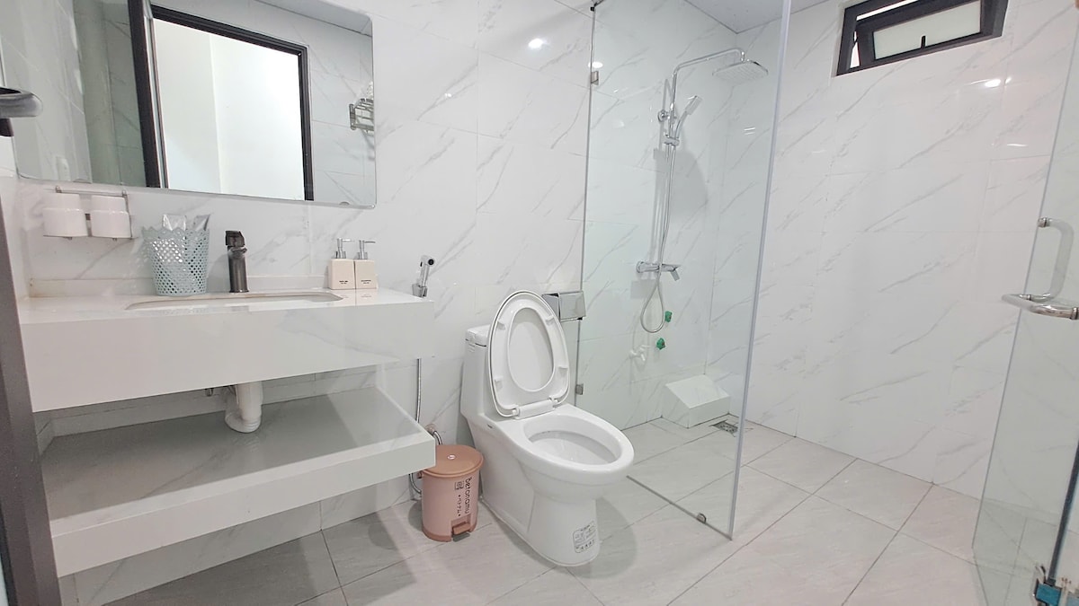 A modern bathroom features a spacious shower area enclosed in glass, complemented by a sleek sink with minimalist storage underneath. A neutral color scheme and contemporary fixtures create a clean and open environment. Natural light filters in through the window, enhancing visibility.