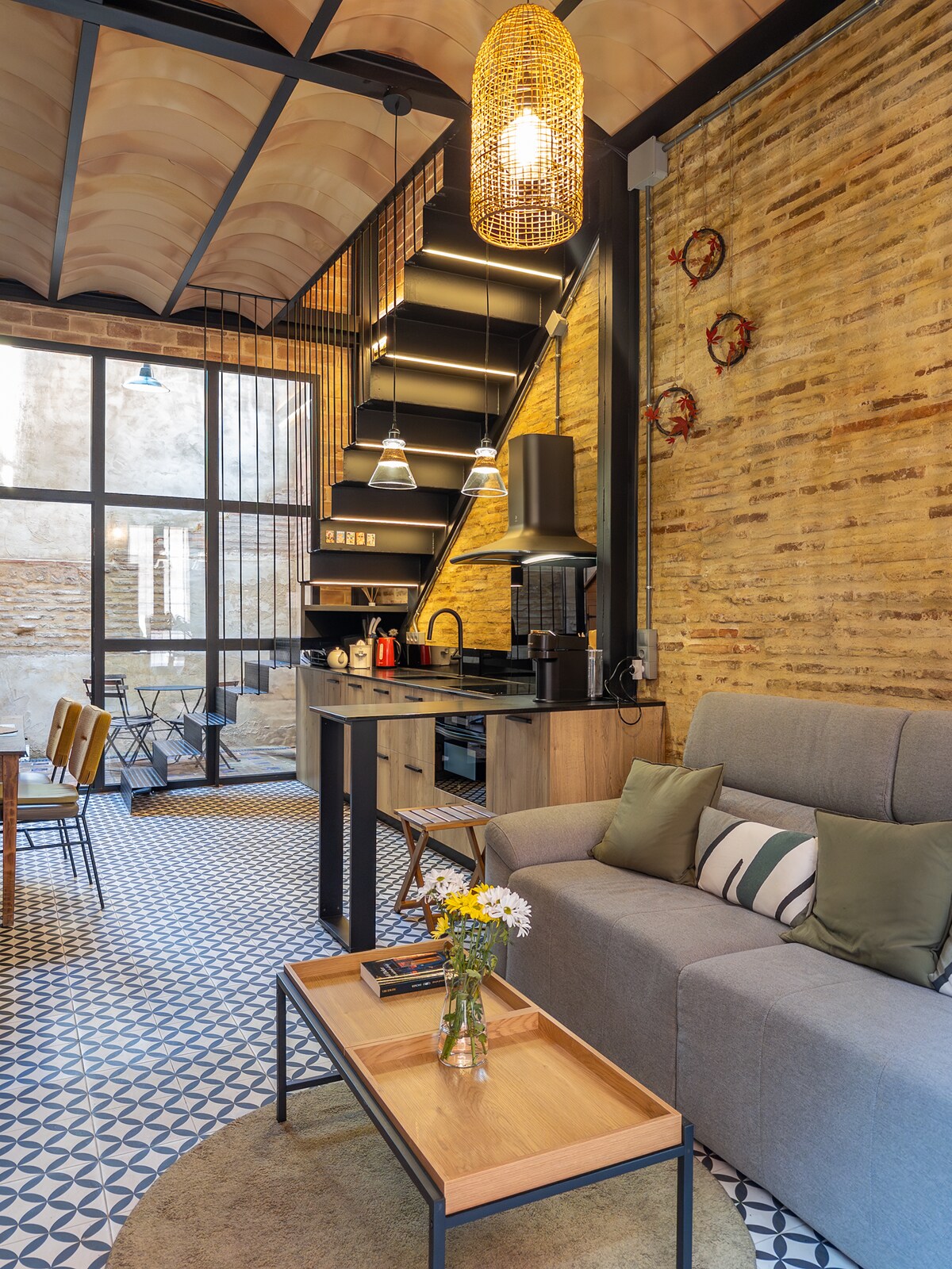 A modern living area features a comfortable gray sofa and a wooden coffee table adorned with a small vase of flowers. A staircase leads to the upper levels, while a kitchenette and dining area are integrated within the open layout, complemented by textured brick walls.