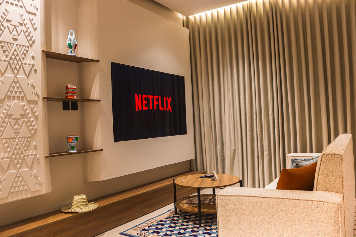 A cozy living area features a light-colored sofa and a round wooden coffee table. A large television mounted on the wall displays the Netflix logo. Textured wall designs and a patterned area rug enhance the layout, with soft lighting illuminating the space.