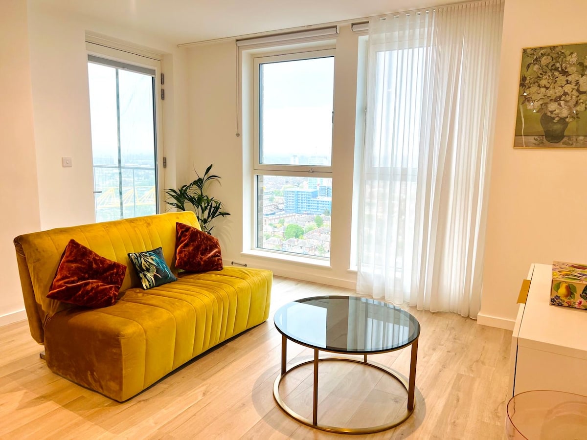 A living space features a yellow sofa with decorative pillows, positioned near large windows that provide natural light and skyline views. A round glass coffee table is placed at the center, complemented by a potted plant in the corner and a wall art piece near the window.