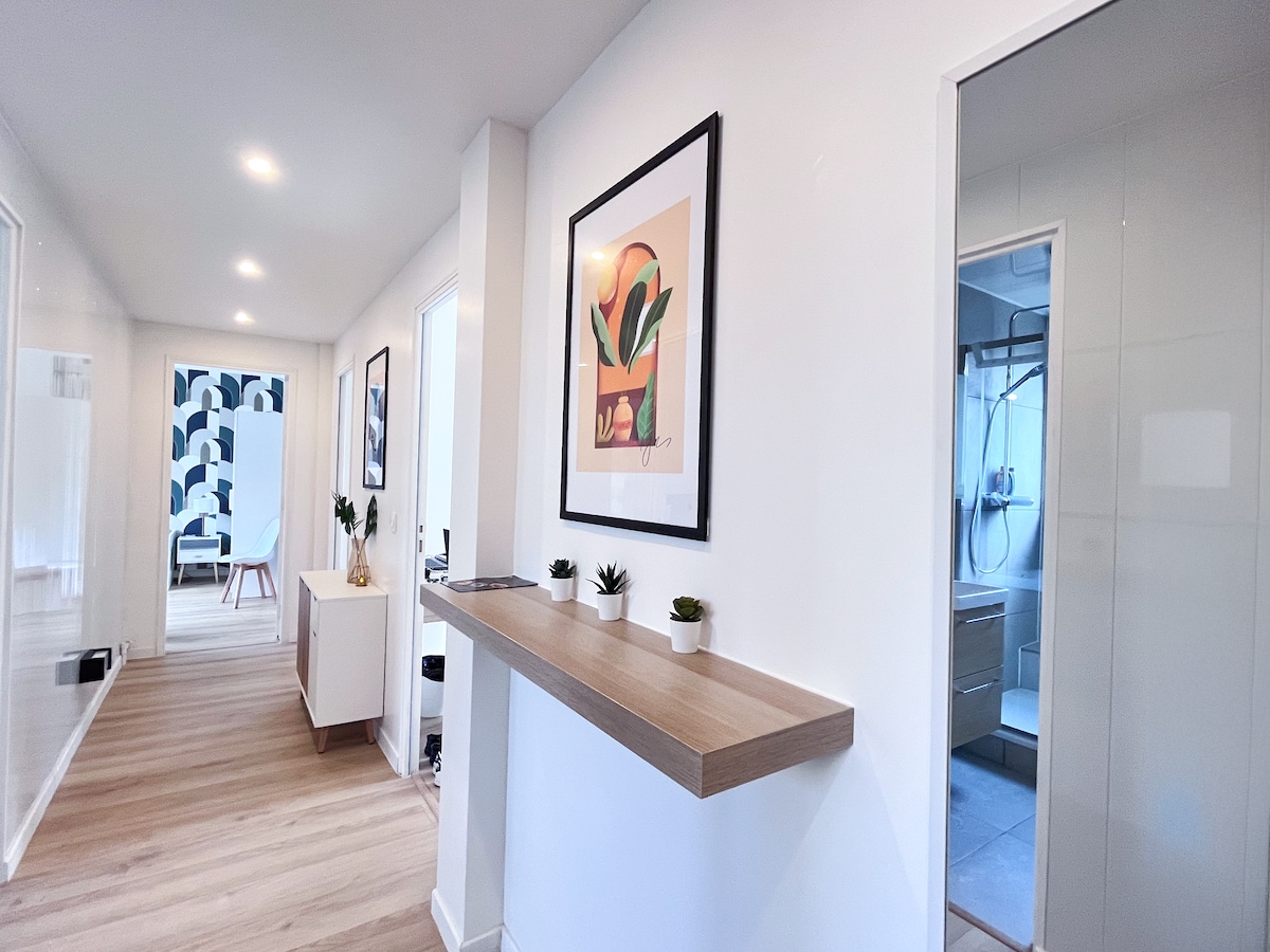 A hallway is shown with light-colored walls and wooden flooring. Original artwork is displayed on the walls, and small potted plants are placed on a narrow wooden shelf. A doorway on the right leads to a bathroom area with modern fixtures.