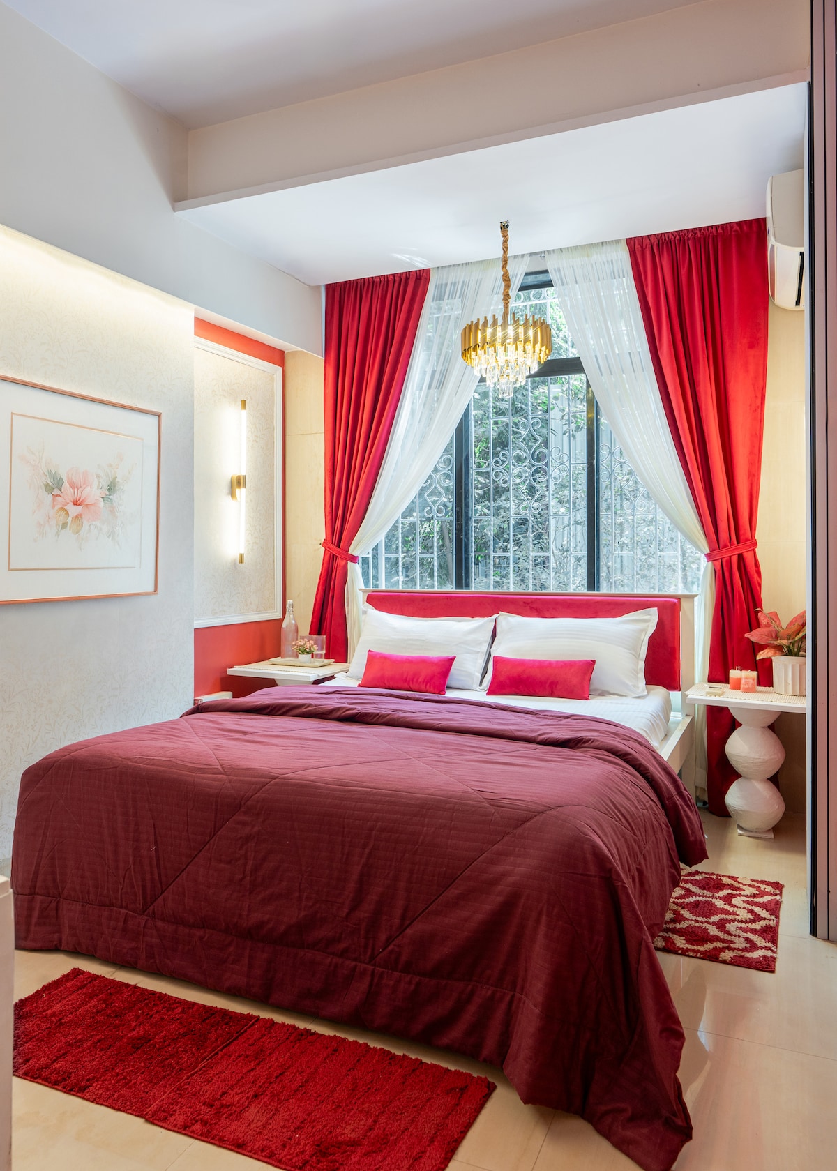 A well-appointed bedroom features a spacious bed adorned with a deep red coverlet and matching decorative pillows. Elegant curtains in red and white frame a window, allowing natural light to fill the space. A small bedside table and a vibrant area rug enhance the inviting atmosphere.