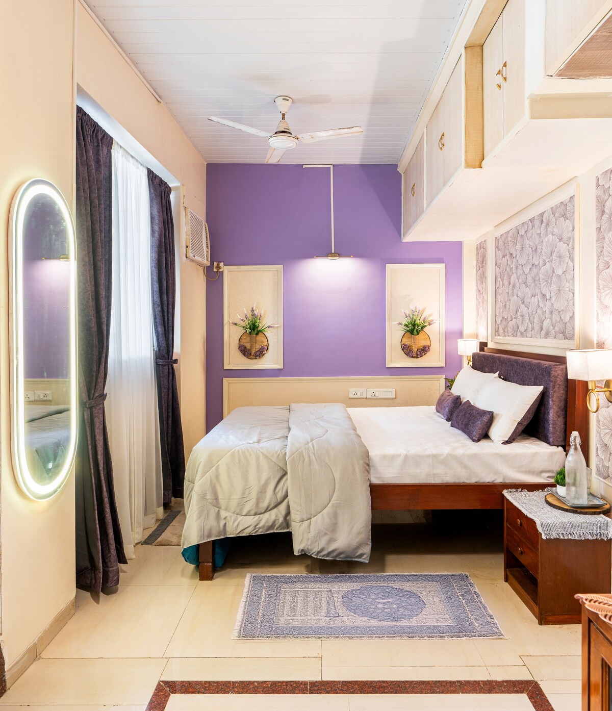 A comfortable bedroom is featured with a spacious bed, accented by soft bedding and decorative pillows. A mirror is mounted on the wall, reflecting the soft lighting. Light-colored walls enhance the ambiance, while a small rug and twin artworks add visual interest.