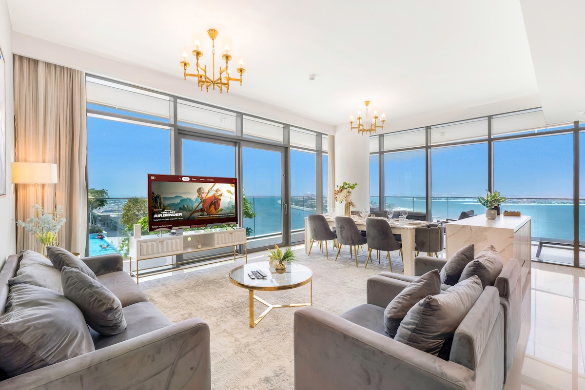 An expansive living area is presented, featuring large glass windows that open to breathtaking sea views. Two grey sofas are positioned around a modern coffee table, while a stylish dining area is visible in the background. A television mounted on the wall adds to the contemporary ambiance.