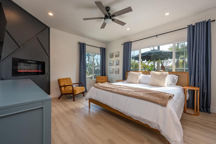 Relájate en esta serena suite king con una pared de chimenea personalizada, sillas de mediados de siglo y luz natural relajante. Rodeado de palmeras y tonos suaves, ¡es un refugio acogedor para relajarse con comodidad y estilo elevado!
