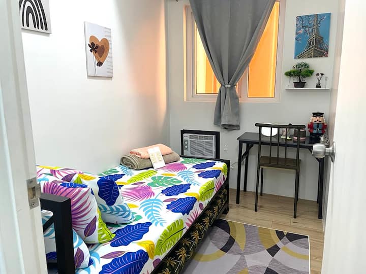 Cozy and vibrant studio with a colorful tropical-themed bed, work desk, and natural light from a curtained window. Features air conditioning, stylish decor, wall art, and essentials for a comfortable stay. Perfect for solo travelers or remote workers