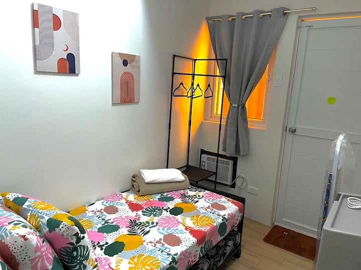 A cozy room with a vibrant single bed, abstract wall art, and soft lighting. It features a clothes rack, gray curtains, a window AC, and a neat white door. Warm yellow light adds a relaxing ambiance to the modern, minimalistic space.