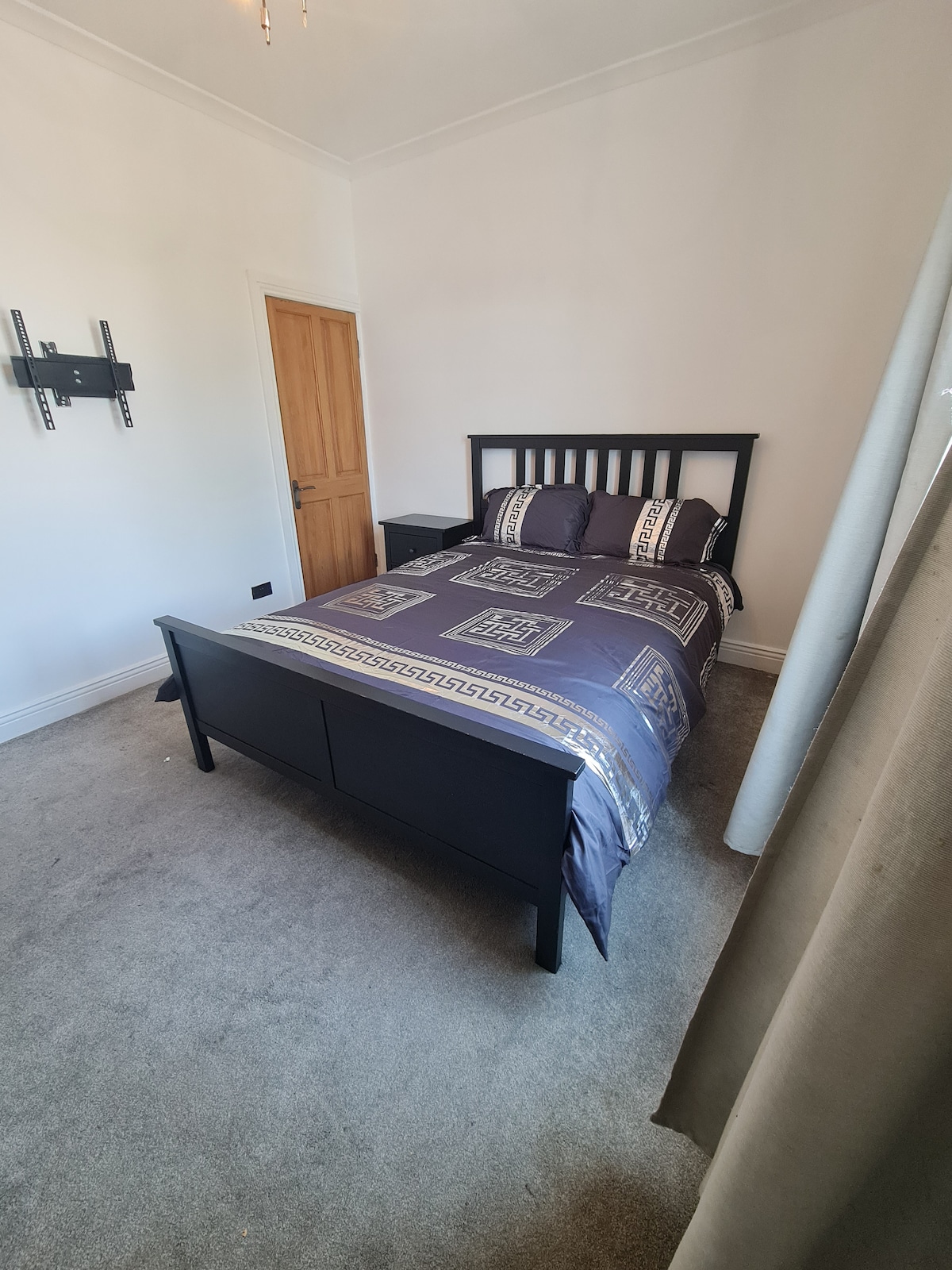 A comfortable bedroom is shown, featuring a plush bed with patterned bedding. A nightstand is positioned beside the bed, and a wall-mounted shelf is visible. A door leads out of the room, with light curtains filtering natural light from a nearby window.