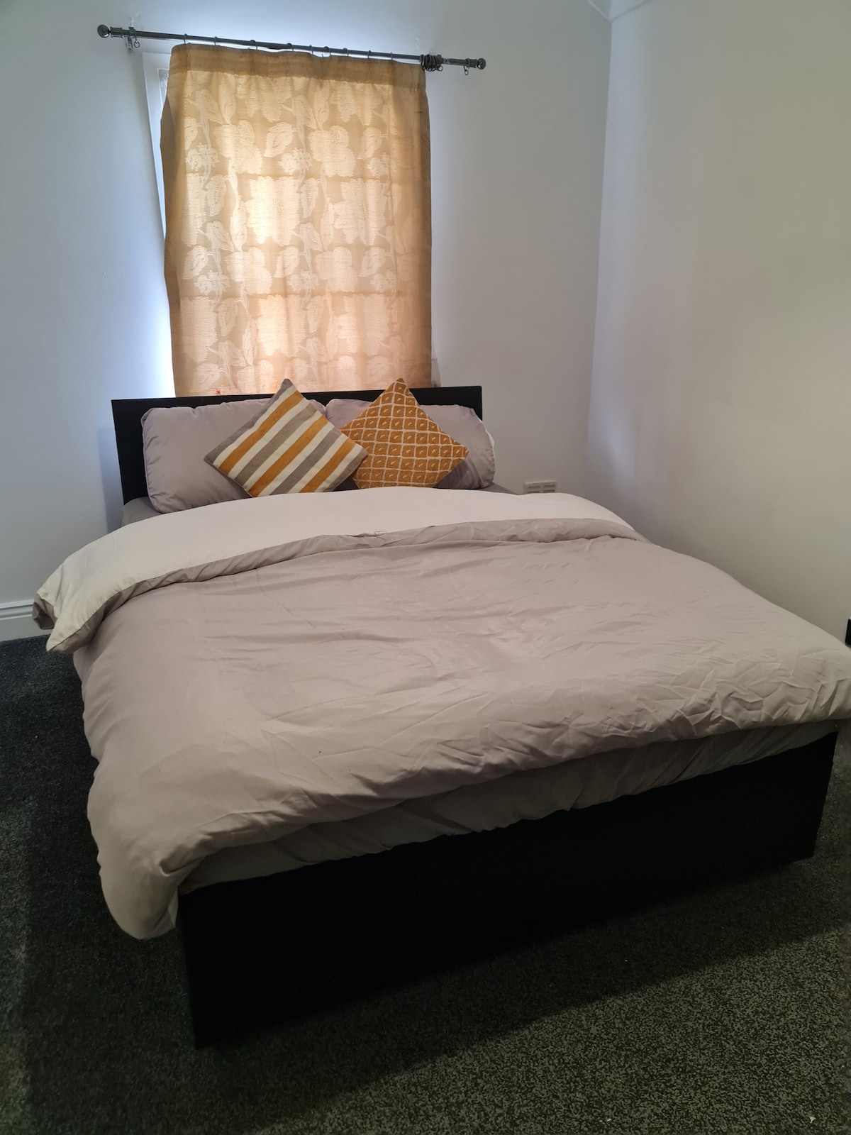 A well-arranged sleeping area features a plush hotel-grade bed dressed in light bedding. Two decorative pillows, one striped and the other geometric, are placed at the head of the bed. Soft, natural light filters through a curtained window, enhancing the calm ambiance.