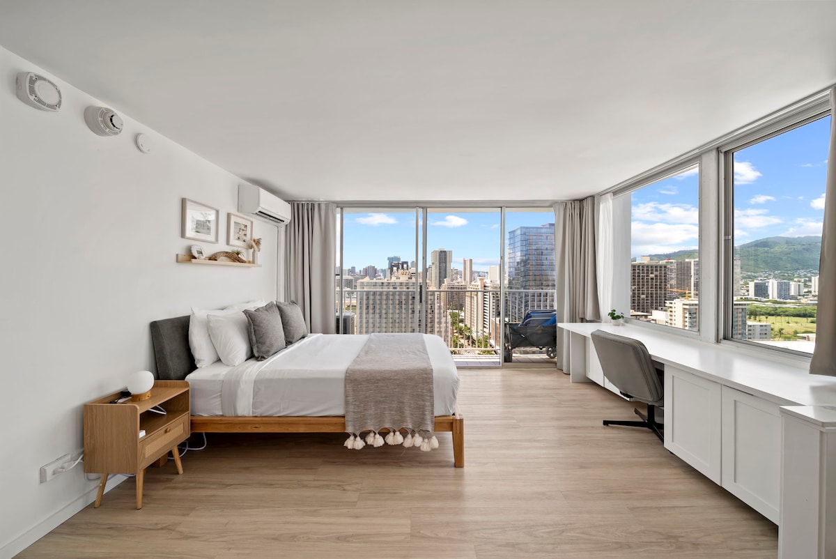 A well-lit bedroom features a spacious bed with neutral bedding, complemented by a decorative throw. Large windows offer panoramic views of the city skyline and mountains. A desk and chair are positioned beside the window, providing a work space with natural light.