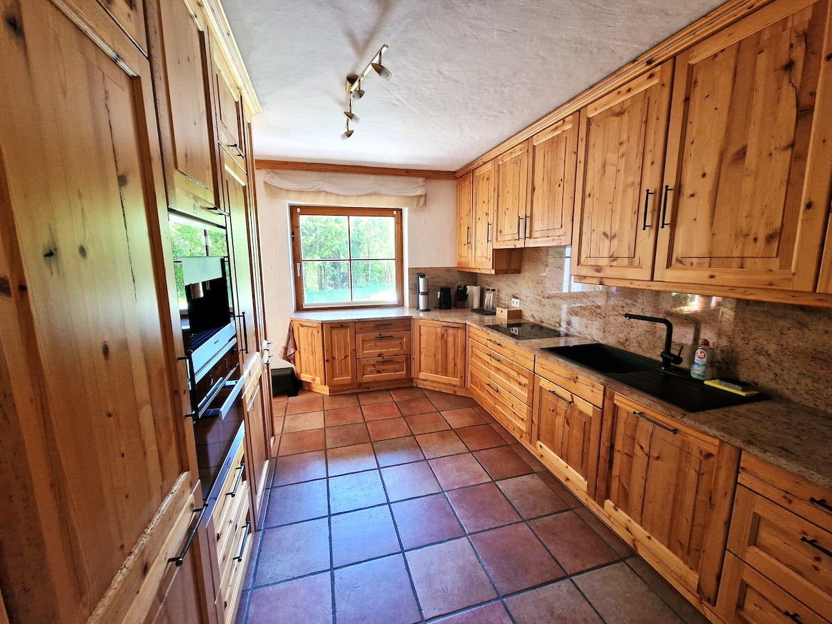 The spacious kitchen features wooden cabinetry and granite countertops. A large window allows natural light to illuminate the space, highlighting a sink and modern appliances. Slate tiles cover the floor, creating a warm and cohesive atmosphere in the heart of the villa.