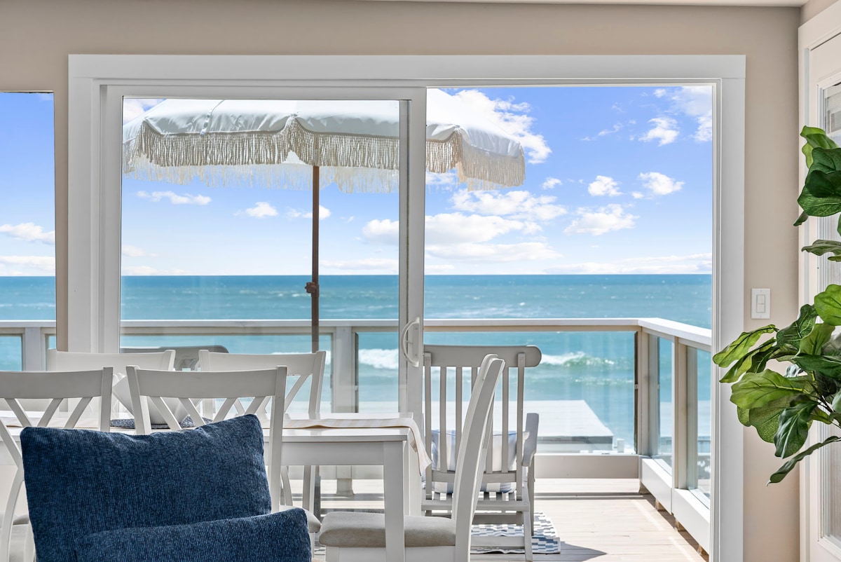 A spacious living area features large glass doors that provide a panoramic view of the ocean. White dining furniture complements the scene, while a sunshade stands ready on the outdoor deck. Soft clouds drift across the blue sky above the shimmering water.